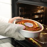 Een persoon met ovenwanten haalt voorzichtig een goudbruine tulbandcake uit een oven met behulp van een bakblik met een verwijderbare bodem.