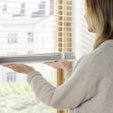 Een vrouw past jaloezieën aan door aan een koord te trekken en zo het licht en de privacy te regelen. Houten lamellen kantelen voor lichte verspreiding.