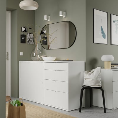 Moderne groene kamer met wit meubilair, waaronder een dressoir, stoel en kruk. Kunstwerken, een spiegel en decoratieve voorwerpen.