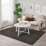 Grijs VINDEBÄK vloerkleed onder beige bank, witte salontafel, kunst aan de muur, plant bij raam.