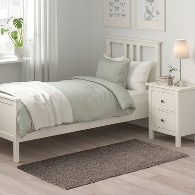 Slaapkamerscène met wit bed, kussens en nachtkastje. Grijs kleed onder bed, achtergrond met planten, lampen en kunst aan de muur.