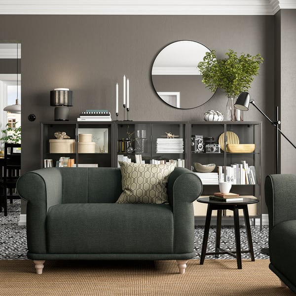 Moderne woonkamer met donkergrijze VISKAFORS fauteuil, geometrisch sierkussen, zwarte planken, decoratieve items en een ronde spiegel.
