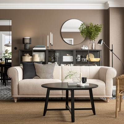 Een gezellige woonkamer met een VISKAFORS beige bank, zwarte salontafel en bruine muur. Op de bank staan zwarte kussens met een patroon.