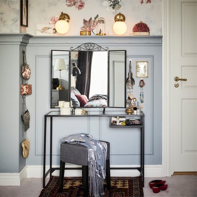 Moderne kaptafel met glazen spiegel, zilveren kruk en grijze tafel met items erop. Grijze muur met wandlampen en bloemendecoraties.