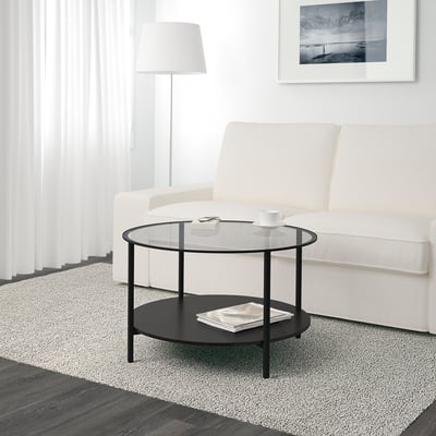 Een moderne VITTSJÖ salontafel met een glazen blad en een zwart metalen frame, met een onderste plank om in op te bergen.