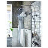 Moderne, zilverkleurige metalen doucheopstelling met wand-, overhead- en handunits. Witte handdoeken, groene plant en een muur met blauw patroon zichtbaar.
