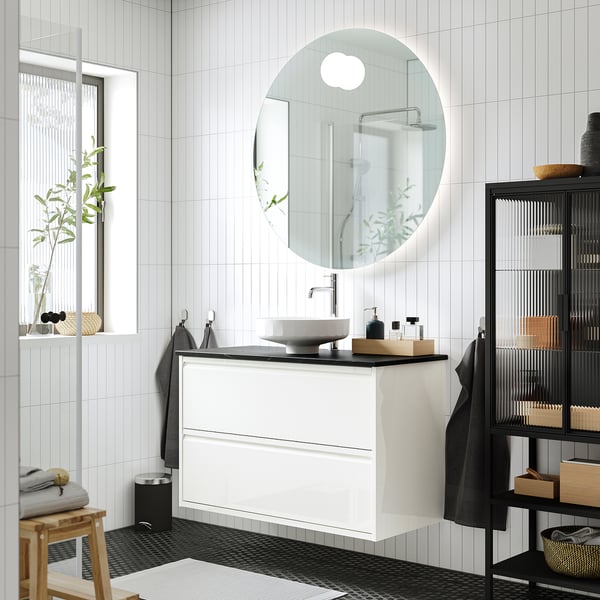 Modern bathroom with ÄNGSJÖN white washstand, round basin, wall-mounted, sleek design, multiple drawers, large mirror, black & white tiles.