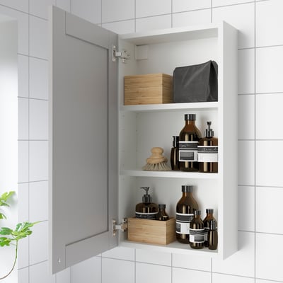 Open bathroom cabinet with shelves storing toiletries and boxes. Grey framed doors, part of ENHET series.