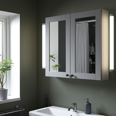Modern grey ENHET mirror cabinet above white sink with lighting, reflecting shower curtain and items, enhancing bathroom brightness.