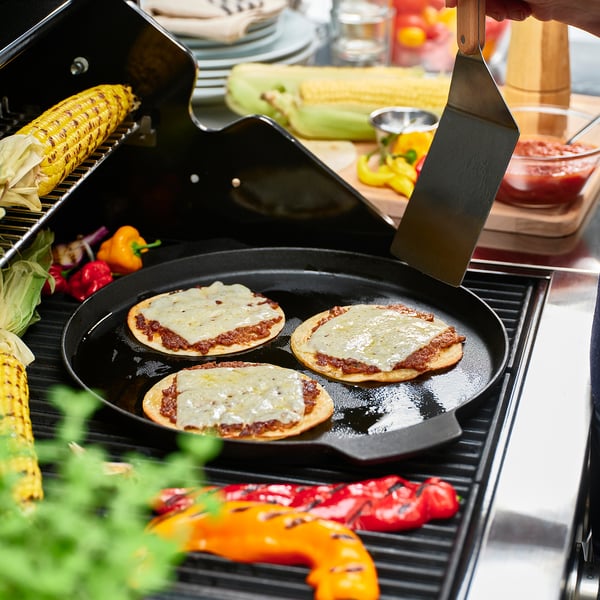 Hand lifts sizzling pizza on cast-iron griddle over bbq, surrounded by veggies.