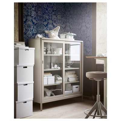 An IDÅSEN cabinet in beige. It has glass sliding doors and four interior shelves. The modern design is sturdy with a metal frame and tempered glass.