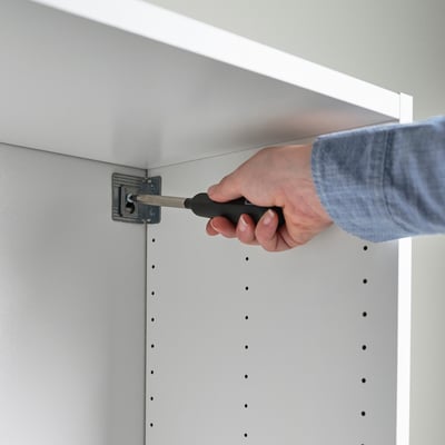 A person is assembling a white cabinet with a screwdriver, securing it to the wall. The cabinet has visible holes for screws.