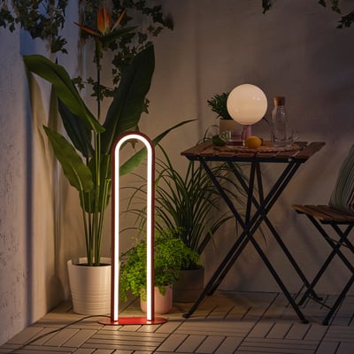 Modern outdoor/indoor lamp, glowing white, stands beside a potted plant.