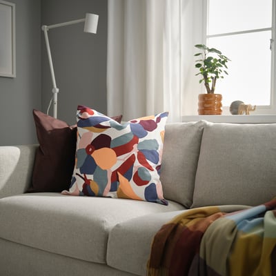 Living room with grey sofa, colourful leaf cushion, dark cushion, folded throw, tall lamp, side plant, sheer curtains.