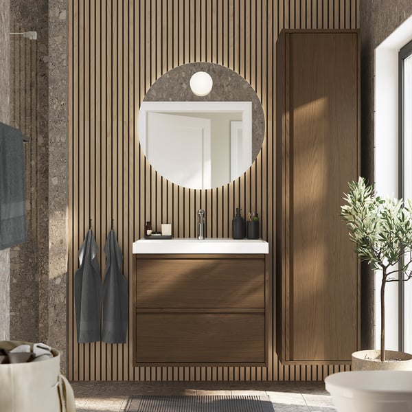 Modern bathroom with large round mirror above wooden cabinet and sink. Dark wood panel walls.