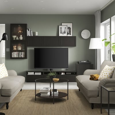 Modern living room with dark green walls, flat screen tv, decorated shelves, grey sofas with yellow pillows, a clock, and a tall lamp.