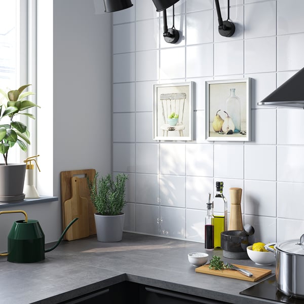 Modern kitchen with white tiled backsplash, hanging lights, plants, art prints, and cooking utensils.