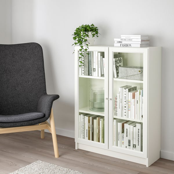 A white BILLY bookcase with glass doors, filled with various books and a plant, standing beside a dark grey chair in a cosy room.