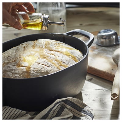 A person drizzles oil from a clear bottle onto flour in a black bowl on a wooden table.