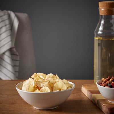 Bowl of chips on wooden table beside bottle and mixed nuts.