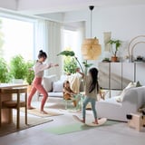 Modern living room with kids playing on green GREJSIMOJS balance board. Light-filled, minimalist decor with plants.