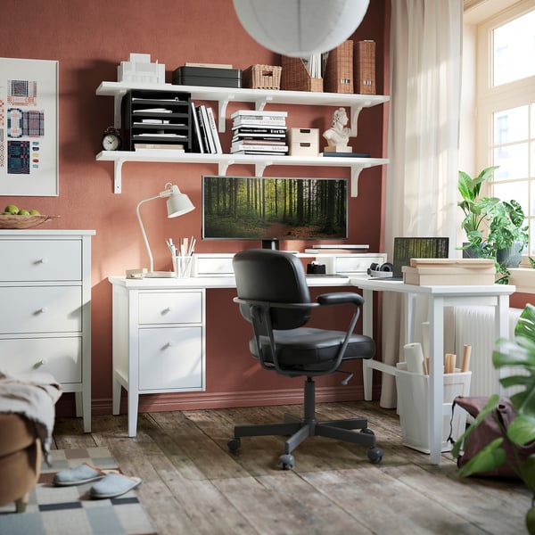Home office setup: l-shaped desk, chair, & decor. White desk with storage, black chair, salmon wall, window, art, & plants.