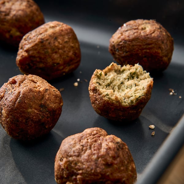 Crispy falafel balls on tray, one cut open revealing internal texture. Onions, chickpeas, peas visible.