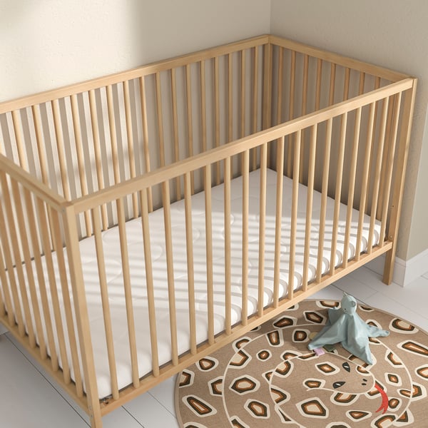 Lightwood KRUMMELUR crib with white mattress, small toy, & patterned rug. Crib & toy small vs. Rug.