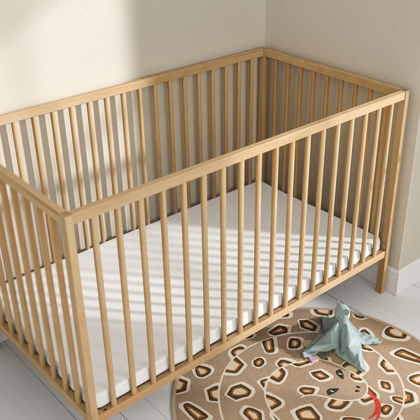 Baby crib with wooden frame, white mattress, and tan rug. Grey cuddly toy nearby. Clean, simple nursery setup.
