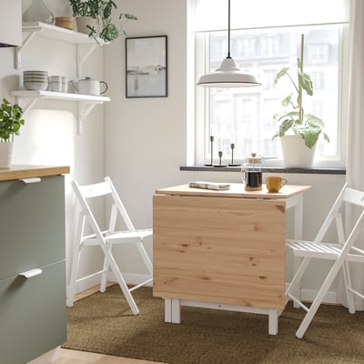 Modern kitchen with a folding light wooden PINNTORP table, white chairs, and house plants. Table expanded, near a green cabinet. Bright, cosy decor.