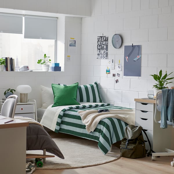 Bedroom with striped green-grey bedding, wood edges, potted plants, desk, and posters. Bright & organised.