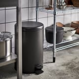 Black kitchen rubbish can with pedal mechanism near wire shelving.