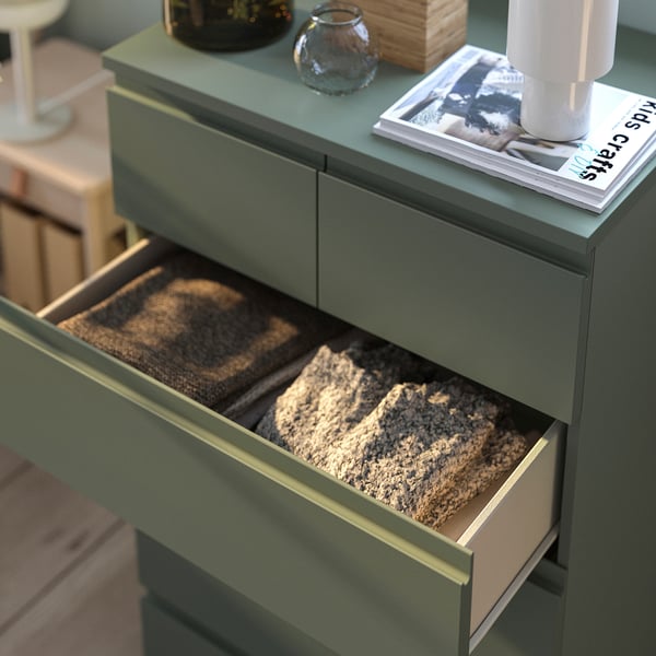 Green STORKLINTA chest with open drawer showing folded blankets. Top holds a magazine, vase, and candle.