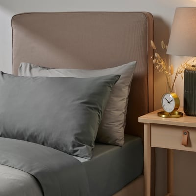 Modern bedroom scene with grey ULLVIDE bedding on large bed; wooden bedside table holds gold clock, green vase with dried flowers, and brown lamp.