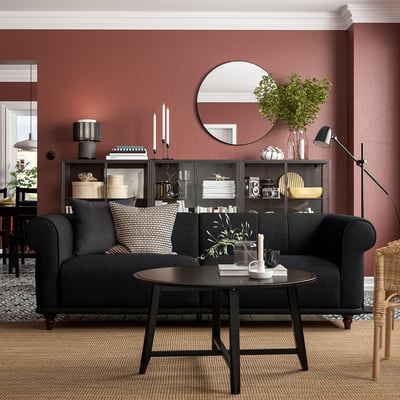 Cosy living room with black l-shaped sofa against red wall, featuring patterned pillows and a round coffee table with a vase and books.