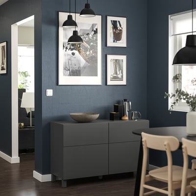 Modern living room with dark blue walls, grey storage unit, wooden chair, and hanging black lamps. Artwork displayed on walls.