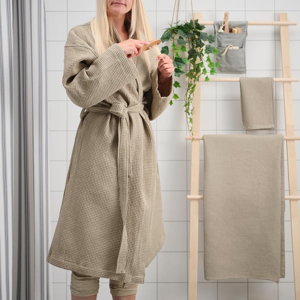 Person in beige robe holding towel, standing by wooden ladder with towels and plant.