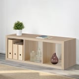 Wooden KALLAX shelving unit with storage boxes, vases, and plant on top, against a wall. Used for display and storage.