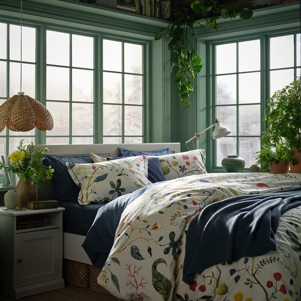 Bedroom with white bed, floral duvet, green cabinet, vase, lamp, views.
