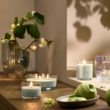 Small LOTSFÅGEL candles of different sizes on table with green fruit and glass vases.