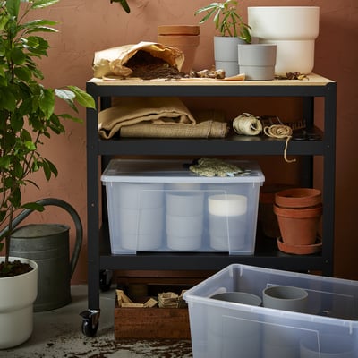 Storage shelf with SAMLA boxes, pots, gardening supplies, and plants against a reddish wall.