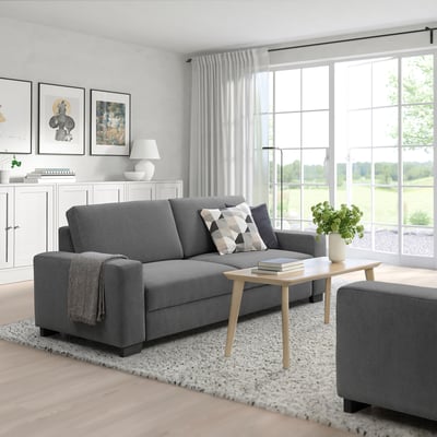 Modern living room with grey SÖRVALLEN sofa, matching ottoman, wooden coffee table, white curtains, and large windows offering a green view.
