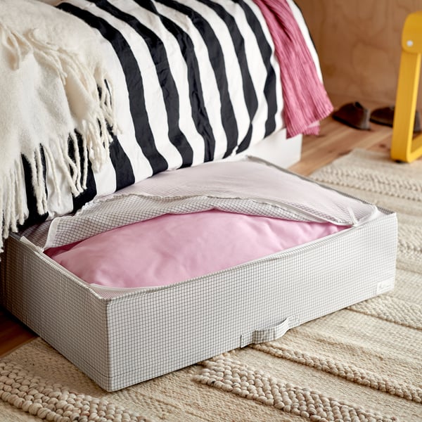Open storage bin under bed, showing light pink blanket inside, with black and white striped pillows on the bed.