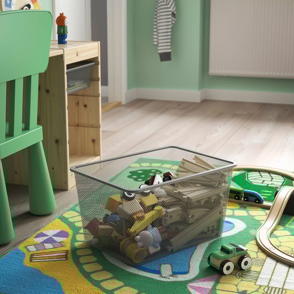 Metal mesh toy basket on colourful rug, holding toys, next to wooden table and green chair in playroom.