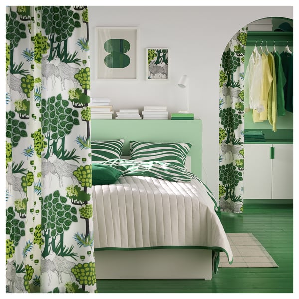 Moderní zelená ložnice se závěsy a ložním prádlem s botanickou tématikou, oddělenou postelí, zeleným kobercem a abstraktními nástěnnými obrazy.