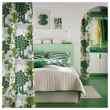 Moderní zelená ložnice se závěsy a ložním prádlem s botanickou tématikou, oddělenou postelí, zeleným kobercem a abstraktními nástěnnými obrazy.