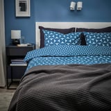 Moderní ložnice s modrým vzorovaným povlečením. Modré stěny, šedý noční stolek s lampou.