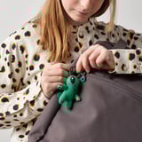 Člověk drží v ruce malou zelenou mimozemskou klíčenku s poutkem na vrchu, určenou k zavěšení nebo připevnění batohu, z kolekce AFTONSPARV.