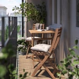 Malý venkovní balkon s hnědým dřevěným stolem a židlemi, polstrovanými sedadly, rostlinami v květináčích a útulným kobercem.