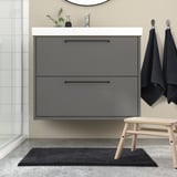 Moderní koupelna s šedou koupelnovou předložkou BACKMÅRA, elegantním toaletním stolkem a ručníky. Podložka se vejde pod umyvadlo s černými akcenty.